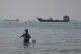 A man stands in the water, appearing to fish, as bulk carriers, cargo ships, and service vessels line the horizon in the Strait of Hormuz off Bandar Abbas, Iran, April 27, 2026