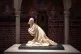 A weathered medieval sculpture of a kneeling man, spotlit on a plinth against the painted brickwork of a castle hall