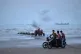 Young men ride motorcycles along the shore as a tractor pulls equipment from the water and commercial vessels appear anchored in the Strait of Hormuz off Bandar Abbas, Iran, July 27, 2026